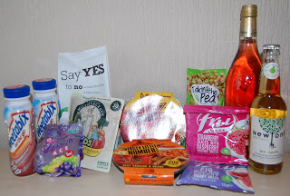 A variety of packaged food and drink items from the July Degustabox, including yoghurt drinks, snack bars, instant noodles, sparkling drinks, crisps, and boxes of snacks, arranged on a flat surface against a neutral background.