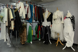 A clothes rail holds various colourful garments, with three mannequins dressed in theatrical costumes—like those seen on a TV show tour such as the Coronation Street Tour—including a white tutu dress, a black and white suit, and white dungarees with a black patterned shirt.