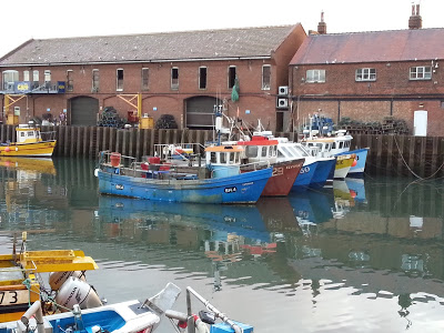 Several small fishing boats are moored in a calm harbour in front of brick buildings, their colourful reflections brightening the water. This tranquil, overcast scene is perfect for a Sunday Photo on your photography blog.