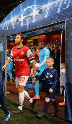 A flashback Friday moment: A footballer in an Arsenal shirt, number 20, walks onto the pitch holding hands with a young mascot, followed by other players, under a UEFA Champions League arch.