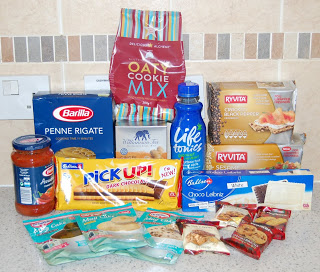 A selection of grocery items from a Degustabox snack subscription box sits on a kitchen worktop, featuring oat biscuit mix, pasta, crispbread, tomato sauce, snack bars, biscuits, chocolate biscuits, crackers, and a blue bottle of drink.