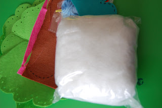 A clear plastic bag filled with white stuffing sits on top of several pieces of colourful felt fabric, including green, pink, blue, and brown—perfect supplies for autumn crafts—on a green surface.