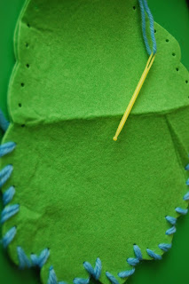 A close-up of a green felt fabric being stitched with light blue wool using a yellow plastic needle, perfect for autumn crafts, against a green background. The edges of the felt have evenly spaced holes for sewing.
