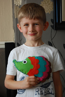 A young boy with light brown hair smiles whilst holding a colourful, felt hedgehog-shaped toy—one of his favourite autumn crafts. He is wearing a white T-shirt with the word "REBEL" printed on it and stands indoors against a patterned wall.