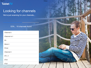 A man in sunglasses and a striped shirt sits on a wooden decking using a tablet. Onscreen text shows a TabletTV review with a channel scan in progress and a list of TV channels found on the left side, highlighting seamless TV streaming.
