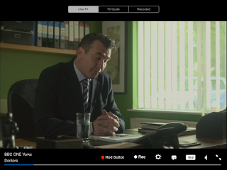 A man in a suit sits at a desk in an office with green walls, looking down thoughtfully. Sunlight streams through vertical blinds. On his desk are a glass of water, files, a phone, and the TabletTV app displaying BBC ONE Yorks Doctors on screen.