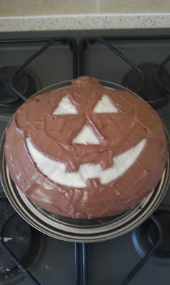 A round cake decorated to look like a jack-o'-lantern, with chocolate icing and white icing forming the eyes, nose, and mouth, sits on a hob—perfect for a Flashback Friday treat!.