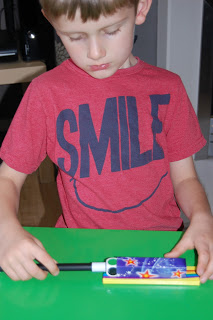 A young boy wearing a red shirt with the word "SMILE" on it is sitting at a green table, looking down whilst holding and examining a colourful rectangular object with stars and a black stick, perfect for a Gee Whizz review.