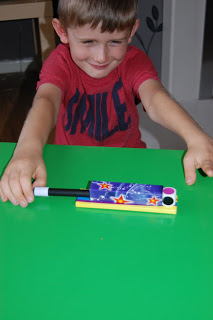 A young boy in a red shirt smiles whilst playing with the colourful Gee Whizz magic trick toy on a bright green table, excited to share his review.