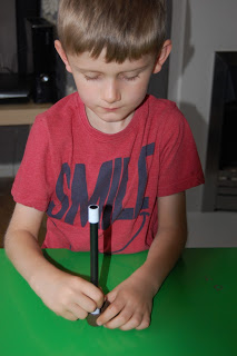 A young boy in a red shirt sits at a green table, holding a black and white marker vertically and pressing its tip onto the table, concentrating on his activity for the Gee Whizz Review.