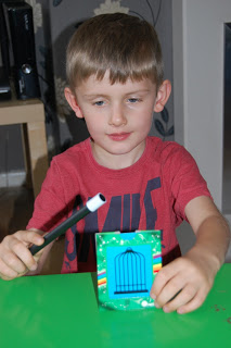 A young boy in a red shirt holds a magic wand and a card with a blue birdcage design, sitting at a green table and looking at the card, ready for his own Gee Whizz review.