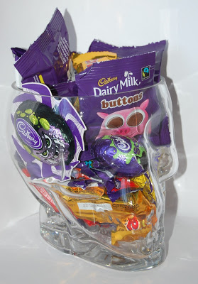 A clear glass skull-shaped container filled with assorted Cadbury chocolates and sweets, including Dairy Milk Buttons and other wrapped treats—perfect for a spooky Halloween treat.