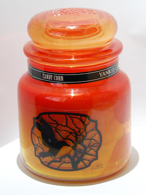 A glass jar candle in orange and yellow shades with a lid, labelled "Candy Corn Yankee Candle." Perfect for Hallowe’en, the jar features a black silhouette of a flying witch on a broomstick with a tree branch design.