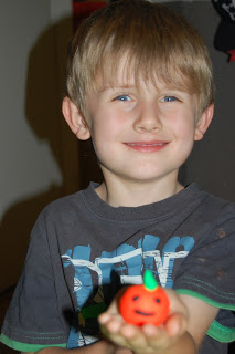 Young boy with blond hair and blue eyes smiles at the camera, holding a small orange clay figure with a green leaf and happy face—perfect for a Flashback Friday moment.