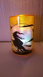 A glowing candle holder features a silhouette of a witch on a broomstick flying in front of a full moon, with clouds in the background. Perfect for Hallowe’en, this Aldi Specialbuys item emits a warm yellow and purple light.