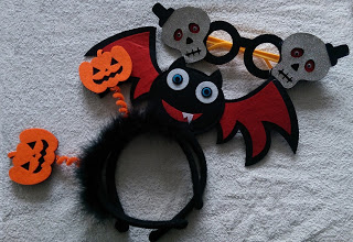 A festive black hairband from Aldi’s Specialbuys, decorated with a smiling bat with red wings, orange jack-o'-lanterns on springs, and spectacles featuring silver skulls, perfect for your Hallowe’en celebrations on a light grey textured surface.
