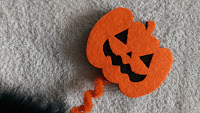 An orange felt jack-o'-lantern decoration with a black mouth and triangular eyes and nose sits on a light grey textured surface. This festive Halloween piece, possibly from Aldi’s Specialbuys, features an orange twisted pipe cleaner stem.