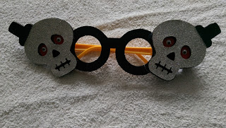 A pair of novelty glasses with black frames featuring two glittery skull shapes on each side, lying on a textured white surface—perfect for Hallowe'en or adding flair to your next Aldi Specialbuys find.