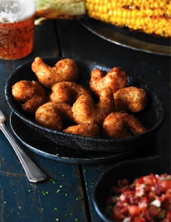 A cast iron frying pan filled with crispy fried prawns sits on a dark wooden table next to a fork, a glass of beer, grilled corn on the cob, and a bowl of salsa—perfect for adding to your list of Halloween recipes.