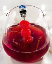 A cocktail with a deep red drink is garnished with three cherries on a skewer topped with a silver skull, surrounded by small white candles—perfect for spooky Hallowe’en Recipes.