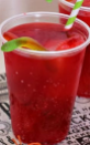 A clear plastic cup filled with a bright red drink, ice cubes, and garnished with a lime slice—perfect for Halloween Recipes. A green-and-white striped straw sits in the drink, with another similar cup partially visible in the background.