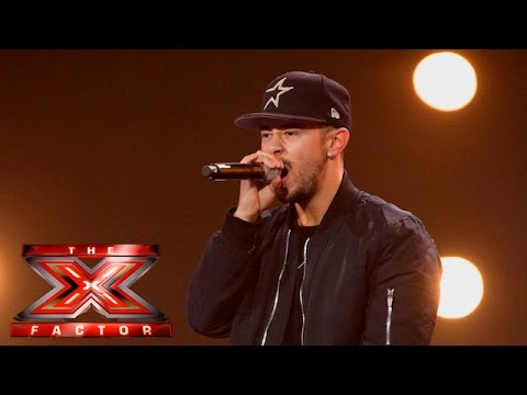 A man wearing a black jacket and black cap sings into a microphone on stage with bright lights behind him, throwing a strop as he passionately delivers his performance. The X Factor logo appears in the bottom left corner.