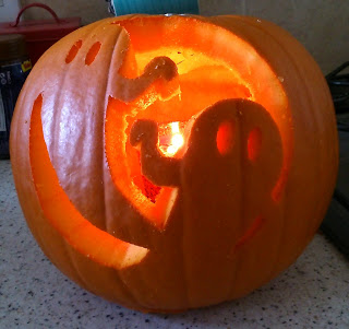 A carved pumpkin with two ghost shapes and a lit candle inside creates a glowing effect on a speckled worktop, making it the perfect flashback Friday centrepiece.