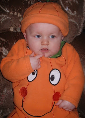 A baby wearing an orange pumpkin costume with a matching hat sits on a patterned brown sofa, touching their chin and looking slightly to the side. This adorable flashback Friday moment features a smiling pumpkin face on the front.