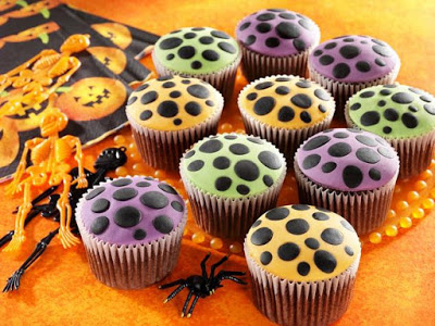 Colourful cupcakes with purple, green, and orange icing, each decorated with black polka dots, are arranged on an orange surface alongside Hallowe’en decorations—perfect inspiration for festive Hallowe’en recipes.