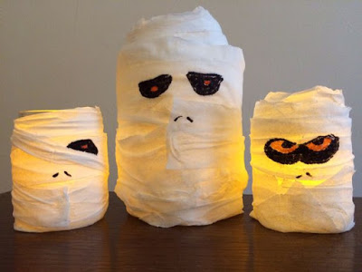 Three glass jars wrapped in white tissue to look like Hallowe’en mummies, with drawn-on eyes and glowing yellow lights inside, sit on a dark surface against a plain background.