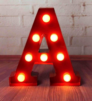A large red Light Up Letter "A" marquee with round bulbs glows brightly, standing on a wooden floor in front of a white brick wall—perfect for your next Giveaway or to Win as an eye-catching decor piece.
