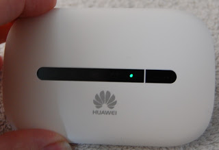 A hand holds a white Huawei E5573 4G Mobile Wi-Fi device with a small green indicator light illuminated on the front.