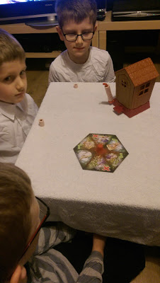 Three boys sit around a table covered with a white cloth, playing Og On The Bog—a board game featuring a hexagonal piece with animal images and a small model house. Perfect for family fun, this review highlights its engaging gameplay.