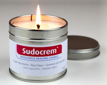 A limited edition Sudocrem Antistress Healing scented candle glows in a metal tin beside its lid on a white surface, with a humorous label listing uses like "Rash Moments - Bad Days - General Grumpiness".
