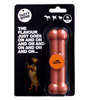 A packaged dog chew toy shaped like a bone labelled "Tasty Bone." The packaging says, "The flavour just goes on and on..." and features a sticker reading "I am bacon," making it one of the best pet gifts with a photo of a dog standing on its hind legs.