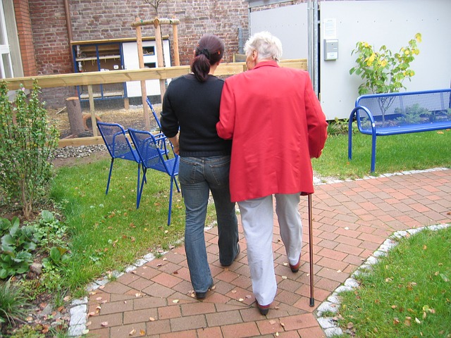 Two people walk arm-in-arm along a brick path in a garden at the care home. One, wearing a red jacket and using a walking stick, is supported by the other in a black top and jeans. Blue benches and green plants surround them.