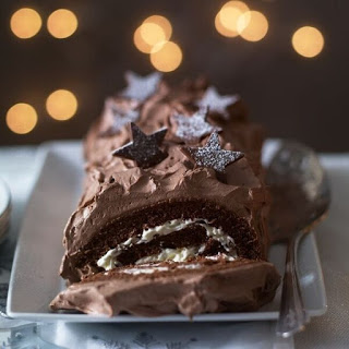 A chocolate Yule log cake—perfect for any chocolate dessert lover—with creamy filling, frosted in rich chocolate, and decorated with sugared star-shaped chocolates. Blurred warm lights glow softly in the background.