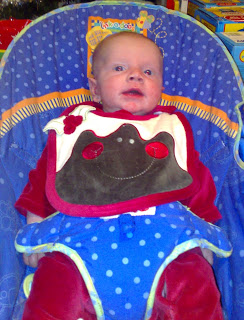 A baby sits in a blue polka-dotted baby chair, wearing a red outfit and a white bib with a brown cupcake design and red buttons, looking slightly upwards—perfect for a sweet Flashback Friday memory.