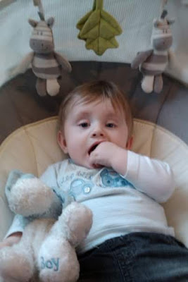 A baby with light brown hair sits in a cushioned seat, sucking on their fingers and holding a white and blue soft toy embroidered with "Boy." Two soft animal toys hang above the baby, making this a perfect Flashback Friday moment.