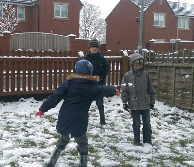 Three children play in a snowy garden beside a wooden fence. One child in the foreground throws a snowball, while the others watch and smile. This joyful MySundayPhoto captures winter fun with houses and trees in the background.