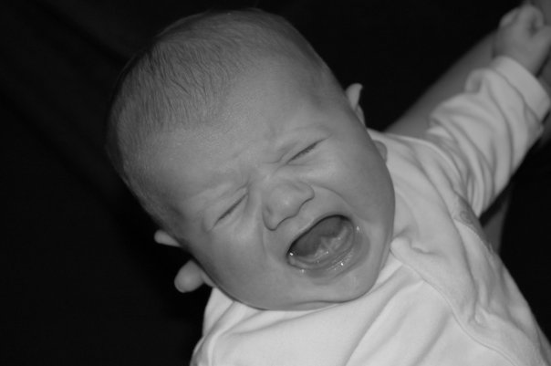 Flashback Friday: Black and white photo of a baby with closed eyes and mouth open wide, appearing to be crying or wailing, wearing a light-coloured shirt against a dark background.