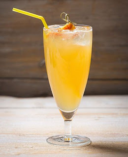 A tall glass filled with a vibrant orange-yellow cocktail, topped with ice, garnished with a slice of fruit, and served with a yellow straw, sits invitingly on a wooden surface.