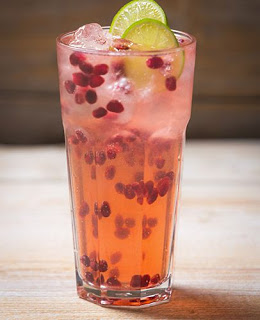 A tall glass filled with a pink cocktail, ice cubes, pomegranate seeds, and two slices of lime on top, placed on a light wooden surface.