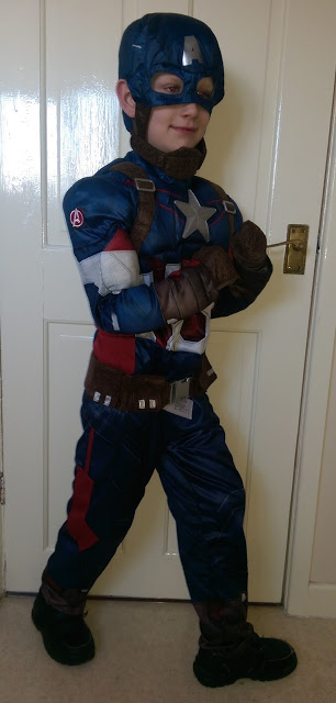 A young boy dressed in a Disney Captain America costume stands sideways and smiles, with one arm bent and holding a door handle, in front of a white door, ready to celebrate World Book Day.