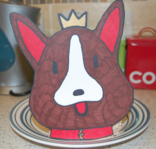A hand-drawn cartoon Corgi cake with a crown and red collar is propped up on a plate in a kitchen setting. The dog themed cake has large ears, a white snout, and its tongue playfully sticking out.
