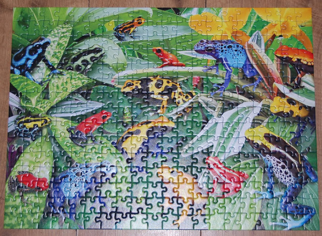 A completed jigsaw puzzle shows a colourful scene full of puzzle fun, with bright blue, red, and yellow frogs sitting on green leaves, surrounded by vibrant foliage and flowers.