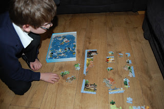 A child with glasses kneels on a wooden floor, enjoying puzzle fun as he assembles pieces of a jigsaw puzzle. The puzzle box and partially completed jigsaw puzzle are in front of him, surrounded by scattered pieces.