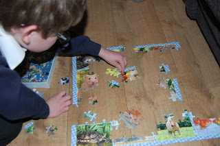 A child with glasses is enjoying Puzzle Fun, assembling a jigsaw puzzle with animal images on a wooden floor. The border is mostly complete as the child carefully fits a piece inside the frame.