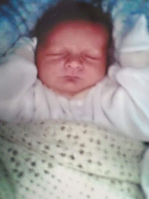 Flashback Friday: A newborn baby is sleeping peacefully with arms raised, wrapped in a soft blanket with star patterns. The baby is wearing a white outfit and resting on a blue, cushioned surface.