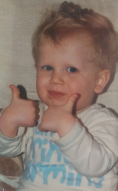 A young child with blonde hair and blue eyes smiles and gives two thumbs up, wearing a white shirt with blue text, sitting on a light-coloured sofa. The perfect Flashback Friday moment!.